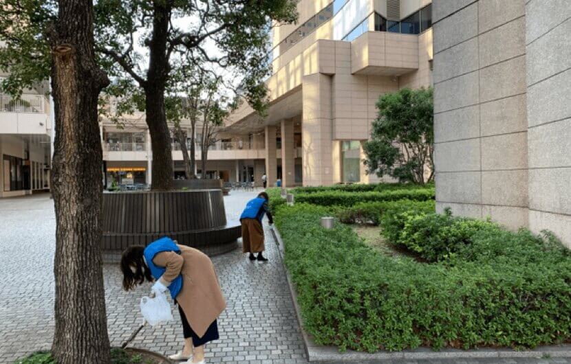 青いベストを着て清掃活動をしている二人の女性。周囲には植え込みや大きな木があり、周囲には建物が立ち並んでいる。
