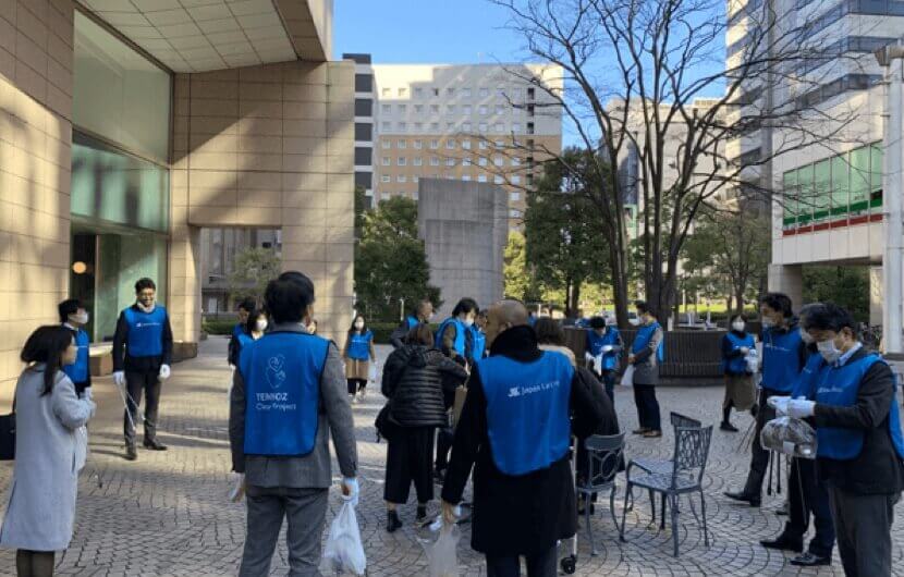 青いベストを着て清掃活動に参加する多数の人々。参加者は各々ゴミ袋を持っており、周囲にはビルや緑の植え込みが見える。